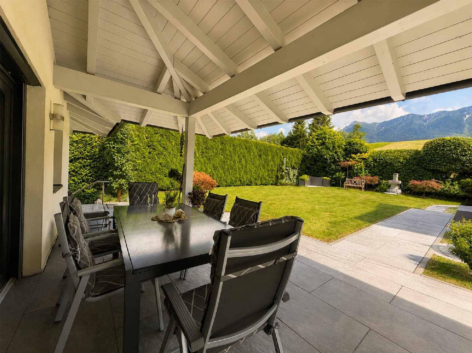 Überdachte Terrasse mit Esstisch und Blick auf den gepflegten Garten und die Berge.
