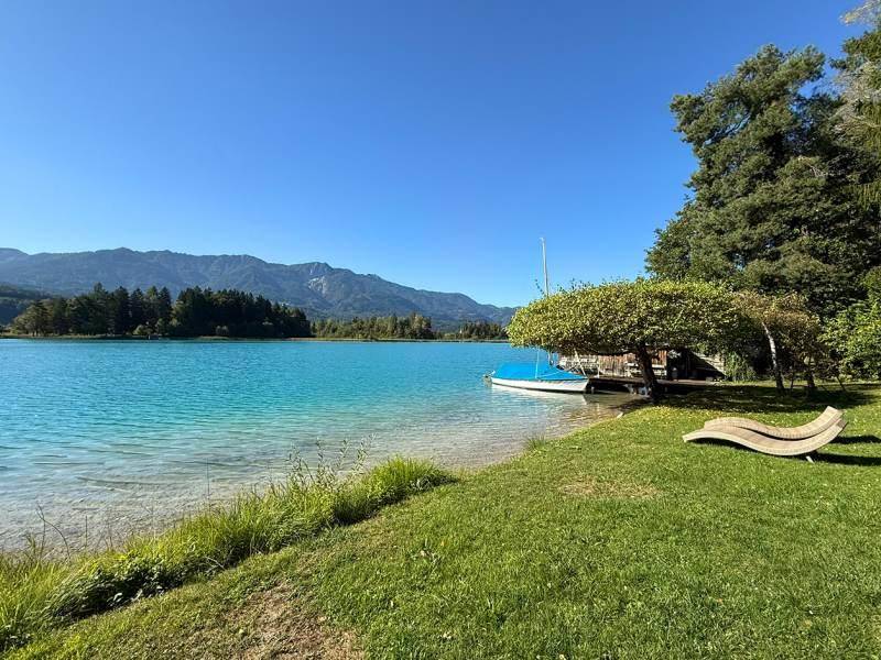 Gepflegte Rasenfläche am See mit Bootssteg und Liegestuhl, umgeben von Bäumen und Bergen.