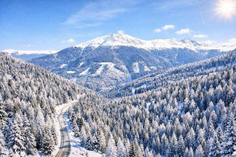 Panoramablick auf eine verschneite Berglandschaft mit einer kurvenreichen Straße und dichten Nadelwäldern.