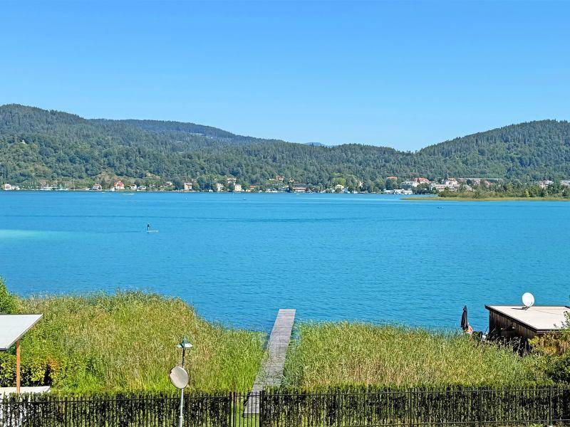 Panoramablick auf den klaren blauen See mit bewaldeten Bergen und einer kleinen Ortschaft am gegenüberliegenden Ufer.
