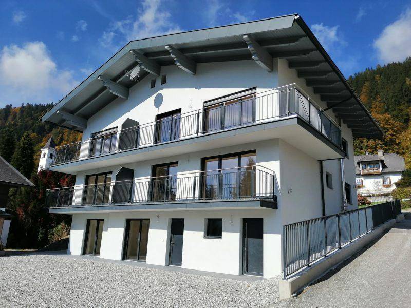 Neubau mit moderner Architektur, mehreren Balkonen und einem Satteldach in bergiger Umgebung.