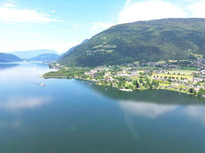 Weitläufiger Blick auf einen großen See mit einer Uferstadt und grünen Bergen unter blauem Himmel.