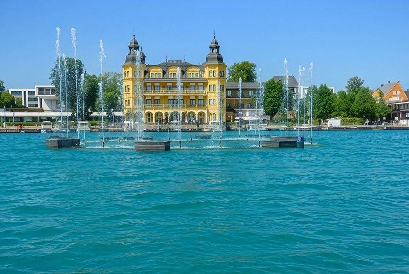 Prächtiges Gebäude am Seeufer mit Wasserspielen und blauer Seeansicht.