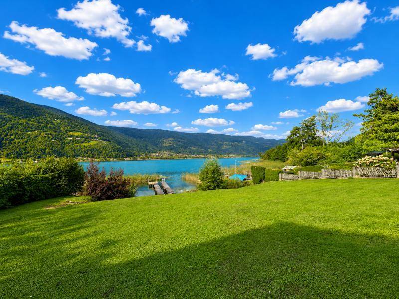 Weitläufige grüne Wiese mit Blick auf den See, einen Holzsteg und die umliegenden Berge.