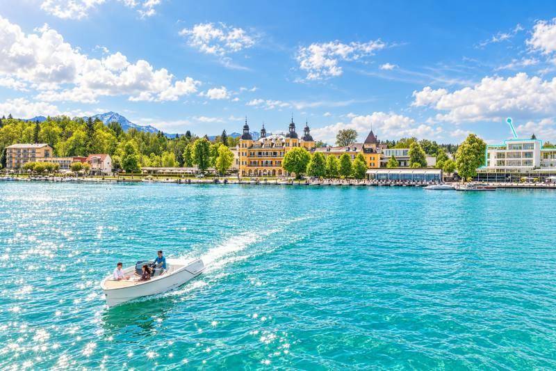 Panoramablick auf den See mit einem Boot, umgeben von grünen Bergen und historischen Gebäuden am Ufer.