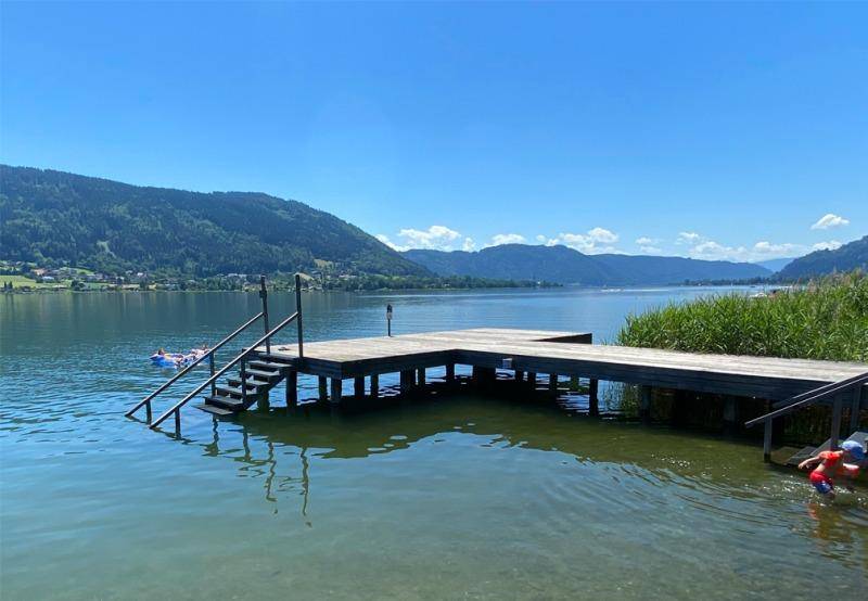 Langer Holzsteg mit Treppen ins klare Seewasser, umgeben von grünen Bergen und Schilf.