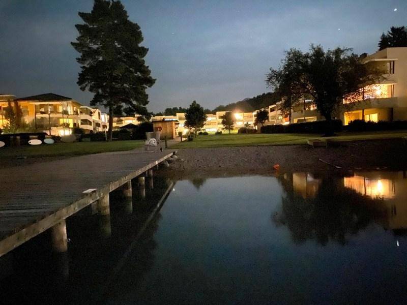 Abendliche Ansicht des Seeufers mit beleuchteten Gebäuden und einem Holzsteg.