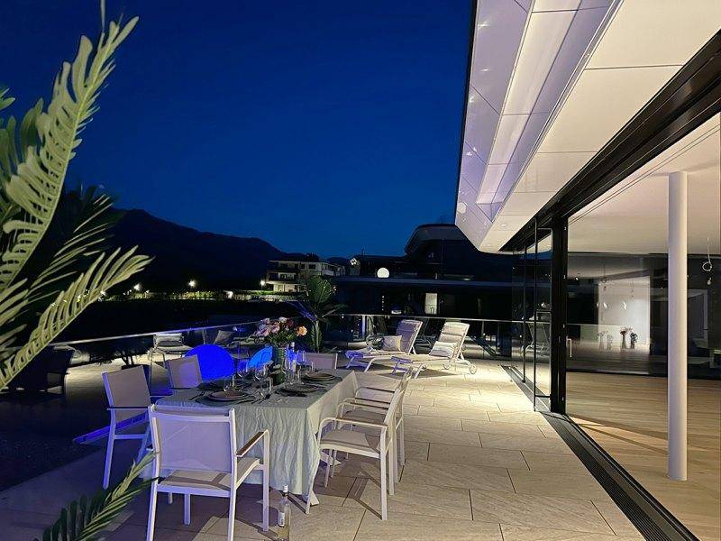 Elegante Außenterrasse am Abend mit gedecktem Esstisch und stimmungsvoller Beleuchtung, Blick auf den See.