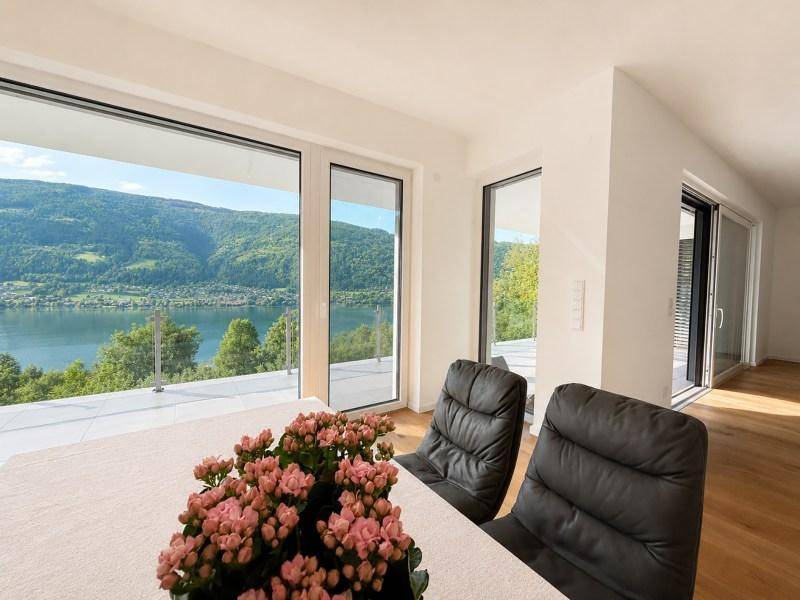 Essbereich mit Seeblick durch große Fenster, ausgestattet mit dunklen Lederstühlen und Blumen.