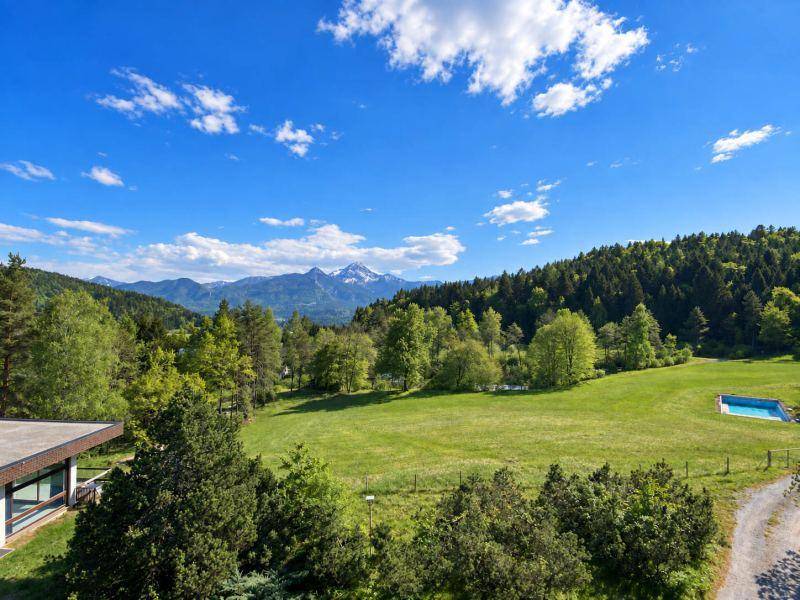 Panoramablick über eine weite grüne Landschaft mit Wäldern, einem Pool und majestätischen Bergen am Horizont.