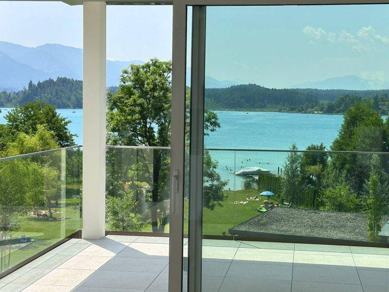Geräumiger Balkon mit Glasgeländer und weitem Blick auf den See, die grüne Landschaft und die Berge.