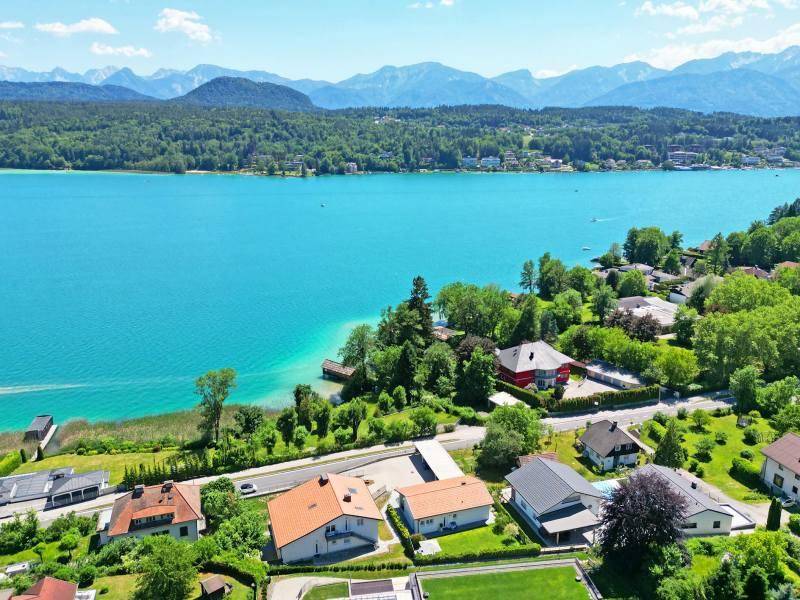 Panoramablick auf eine Wohnsiedlung am Seeufer mit Bergen im Hintergrund unter blauem Himmel.