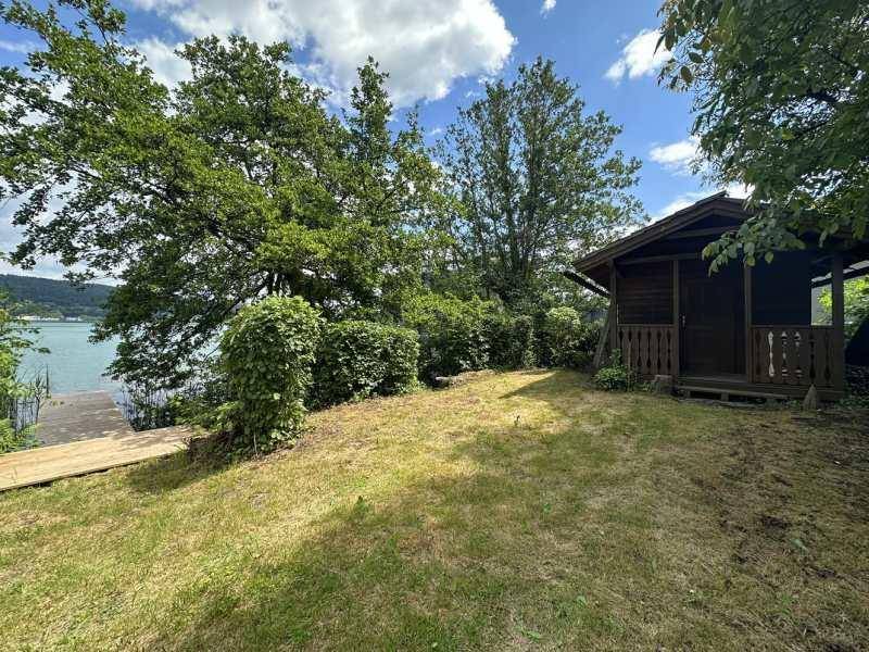 Gartenbereich mit Rasen, Bäumen und einem kleinen Holzhaus direkt am Seeufer.