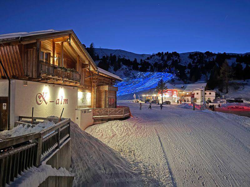 Beleuchtetes Skigebäude "X-alm" in der Nacht, umgeben von Schnee und einer blau beleuchteten Piste.