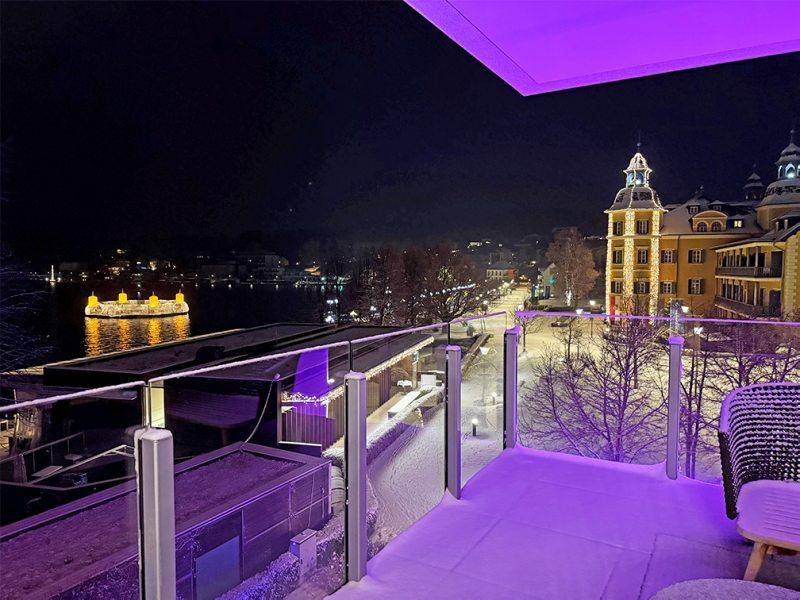 Winterlicher Balkon bei Nacht mit Schnee und Blick auf die beleuchtete Uferpromenade.