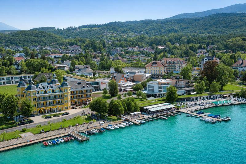 Panoramablick auf das Hotelgebäude, den See und die umliegende grüne Landschaft mit Bergen.