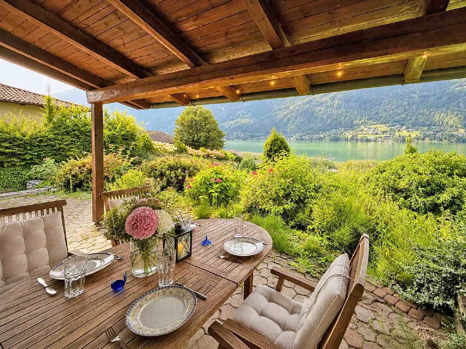 Überdachte Terrasse mit Holztisch und Stühlen, umgeben von üppigem Grün und Seeblick.