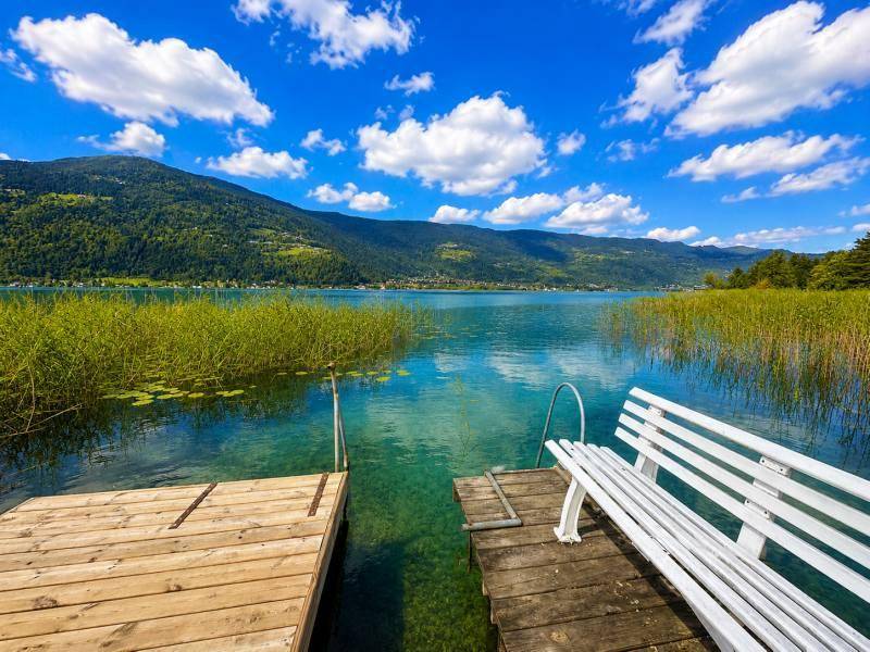 Holzsteg mit weißer Bank am Ufer eines klaren Sees, umgeben von Schilf und Bergen.