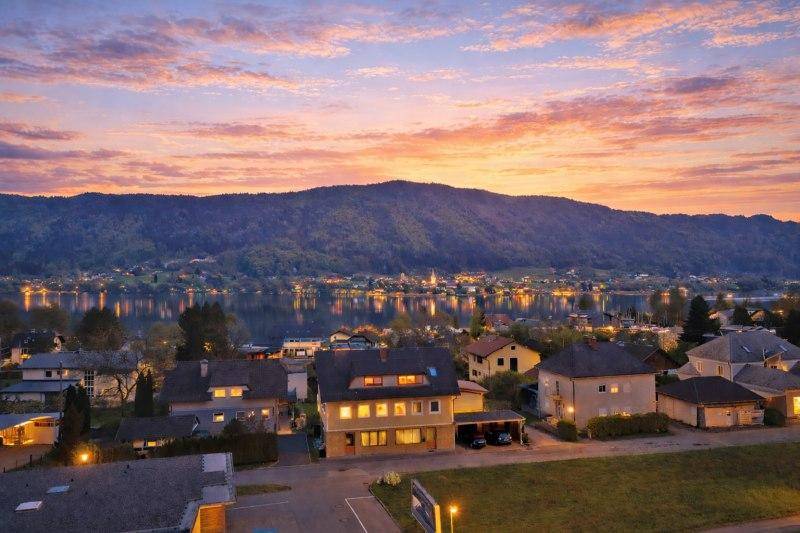 Panoramablick auf eine Stadt am Seeufer bei Sonnenuntergang, umgeben von Bergen und leuchtenden Lichtern.