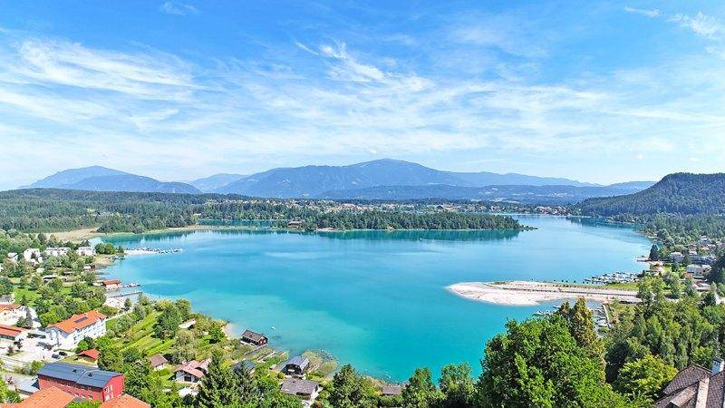 Panoramablick auf einen türkisfarbenen See, umgeben von grünen Wäldern und Bergen unter blauem Himmel.