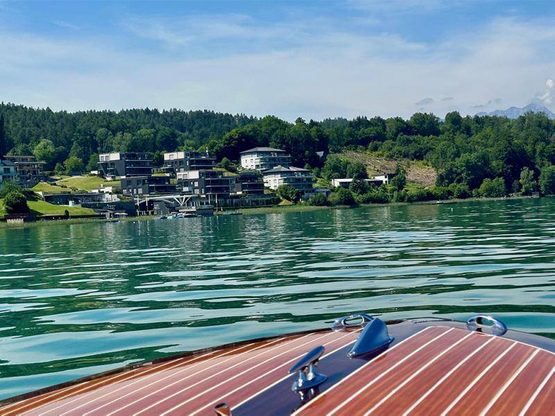 Blick vom See auf die moderne Architektur der Häuser am Ufer und die bewaldeten Hügel.