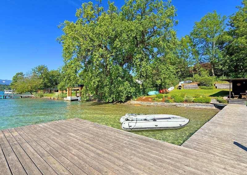 Breiter Holzsteg am Ufer eines klaren Sees mit einem Schlauchboot und üppiger Vegetation.