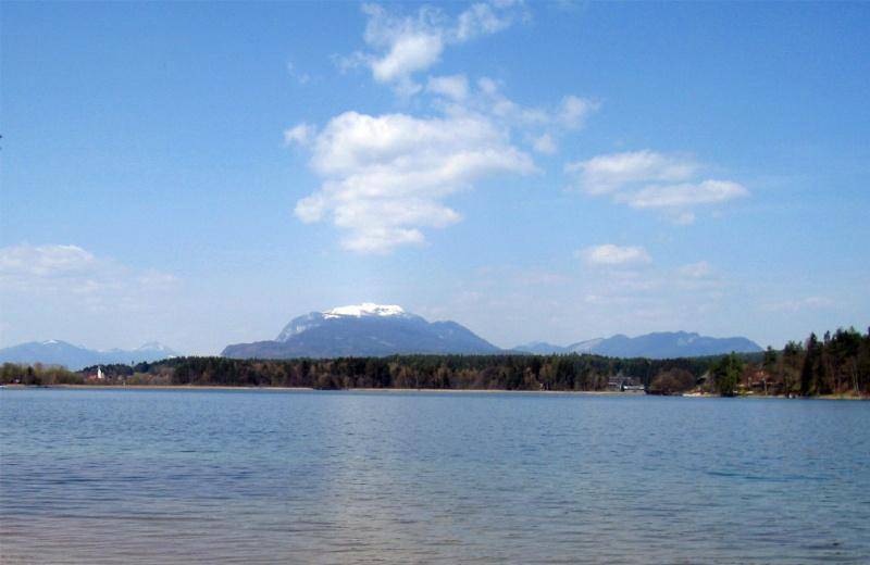 Weitläufiger Blick über den ruhigen See mit schneebedeckten Bergen im Hintergrund unter blauem Himmel.