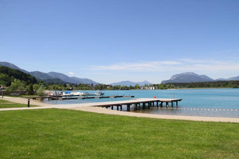 Weitläufiger Seezugang mit Sandstrand, Holzsteg und Bootsanlegestellen vor Bergkulisse.