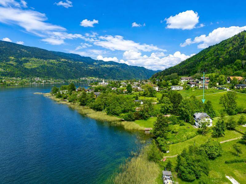 Weitläufige Luftaufnahme der Seenlandschaft mit einem Dorf und bewaldeten Bergen im Hintergrund.