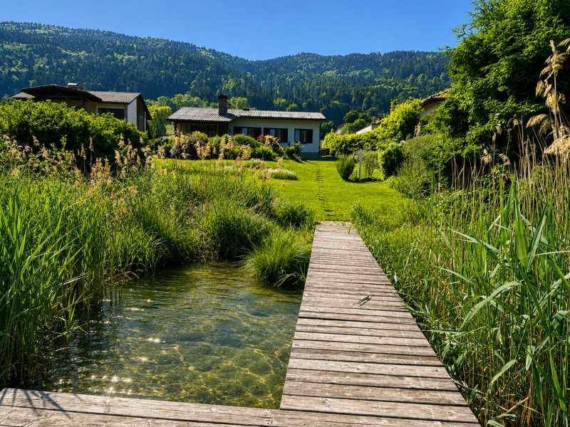 Langer Holzsteg über das Wasser, der zu einem Haus am Seeufer mit üppigem Grün führt.