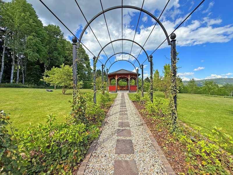 Geschwungener Gartenweg unter einer Pergola, der zu einem charmanten roten Pavillon führt.