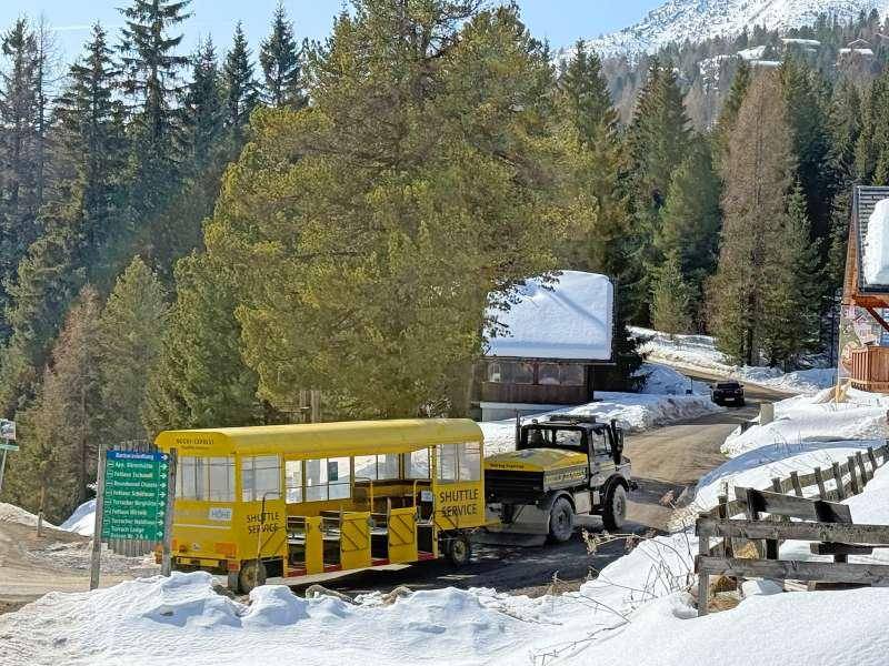 Ein gelber Shuttle-Service-Bus auf einer verschneiten Bergstraße, umgeben von hohen Bäumen und winterlicher Landschaft.