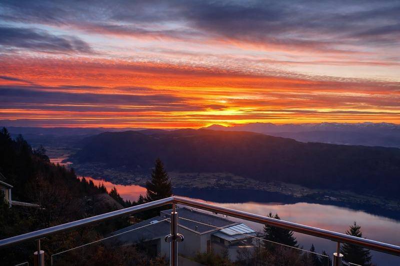 Atemberaubender Sonnenuntergang über einem See und den Bergen, gesehen von einem Balkon mit Glasgeländer.
