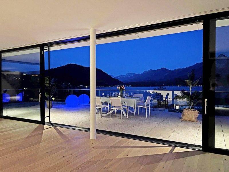 Innenansicht eines Wohnbereichs mit großen Schiebetüren, die sich zu einer beleuchteten Terrasse mit Seeblick öffnen.