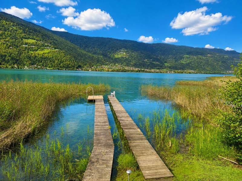 Zwei Holzstege führen in den klaren See, umgeben von Schilf und einer malerischen Berglandschaft.