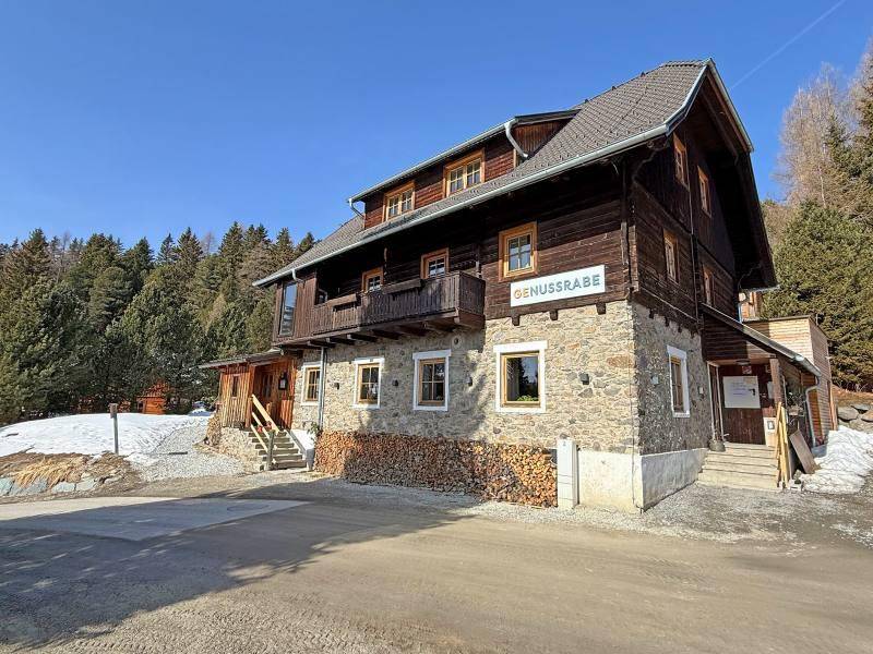 Rustikales Gebäude mit einer Kombination aus Stein- und Holzfassade, umgeben von Bäumen und etwas Schnee am Boden.