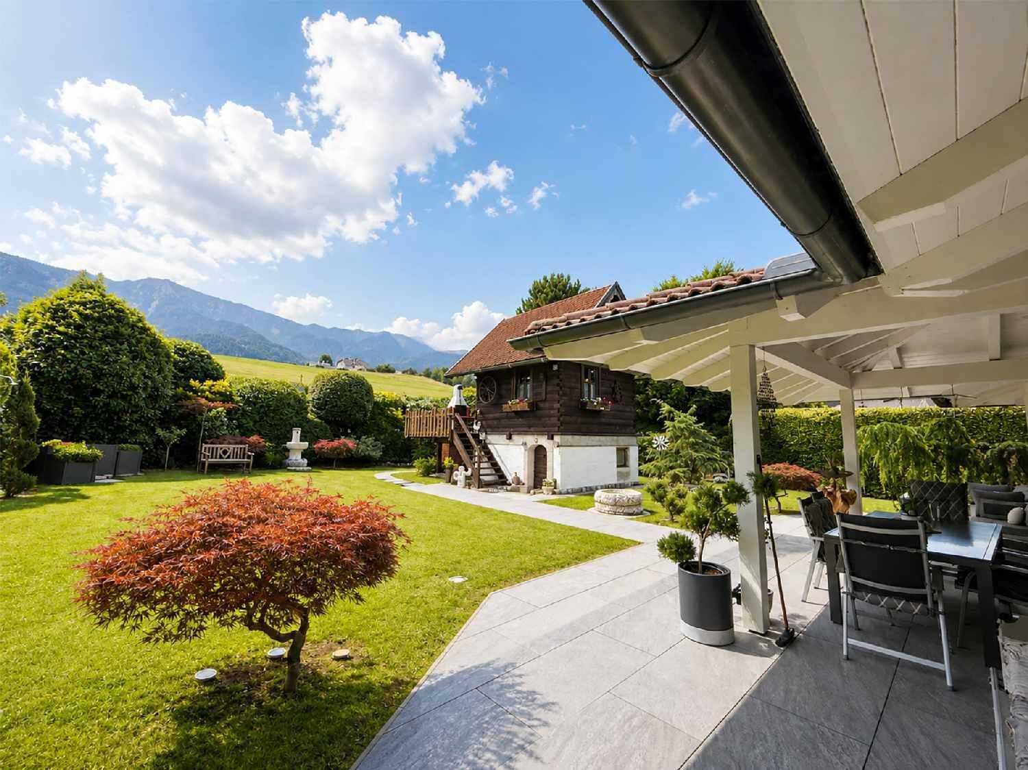 Malerischer Garten mit Terrasse, Holzhaus und Bergblick unter blauem Himmel.