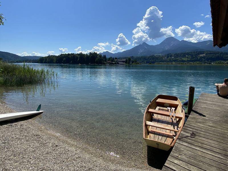 Klarer See mit kleinem Ruderboot am Ufer und Holzsteg unter strahlend blauem Himmel.