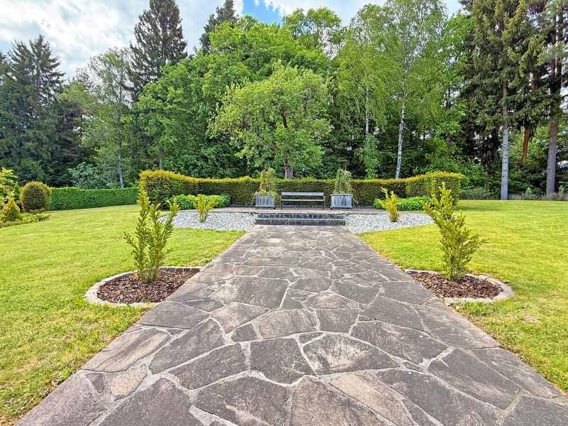 Geschwungener Steinweg führt durch den gepflegten Garten zu einer Sitzbank mit Hecke im Hintergrund.