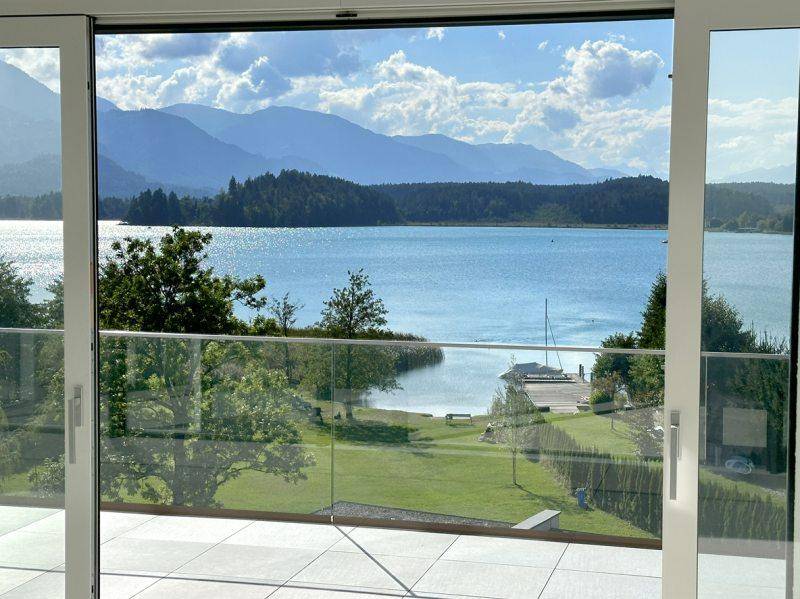 Geräumiger Balkon mit Glasgeländer und weitem Blick auf den See, die grüne Landschaft und die Berge.