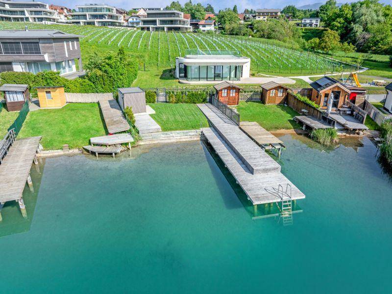 Luftaufnahme eines Seegrundstücks mit Holzstegen, grünen Rasenflächen und modernen Gebäuden im Hintergrund.