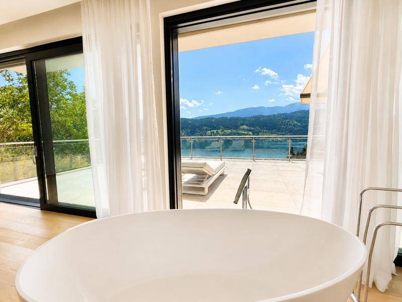 Freistehende Badewanne mit direktem Blick auf den See und die umliegende Berglandschaft durch große Fenster.