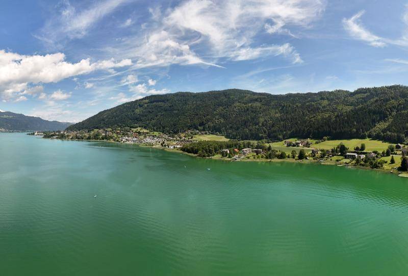 Panoramablick auf den weitläufigen See, umgeben von bewaldeten Hügeln und kleinen Siedlungen unter blauem Himmel.