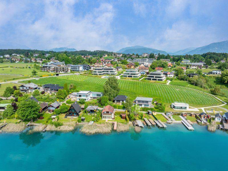 Panoramablick aus der Vogelperspektive auf eine Siedlung am Seeufer mit grünen Hügeln und Bergen.