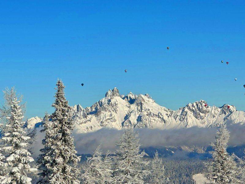 Spektakulärer Blick auf schneebedeckte Berggipfel mit mehreren bunten Heißluftballons am strahlend blauen Himmel.