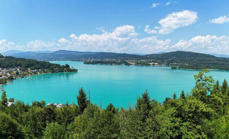 Panoramablick auf den türkisfarbenen See, umgeben von grünen Hügeln und Wäldern unter blauem Himmel.