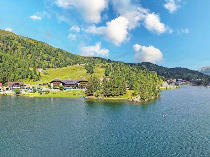 Weitläufiger Blick auf einen See mit Gebäuden am Ufer, umgeben von Bäumen und Bergen.