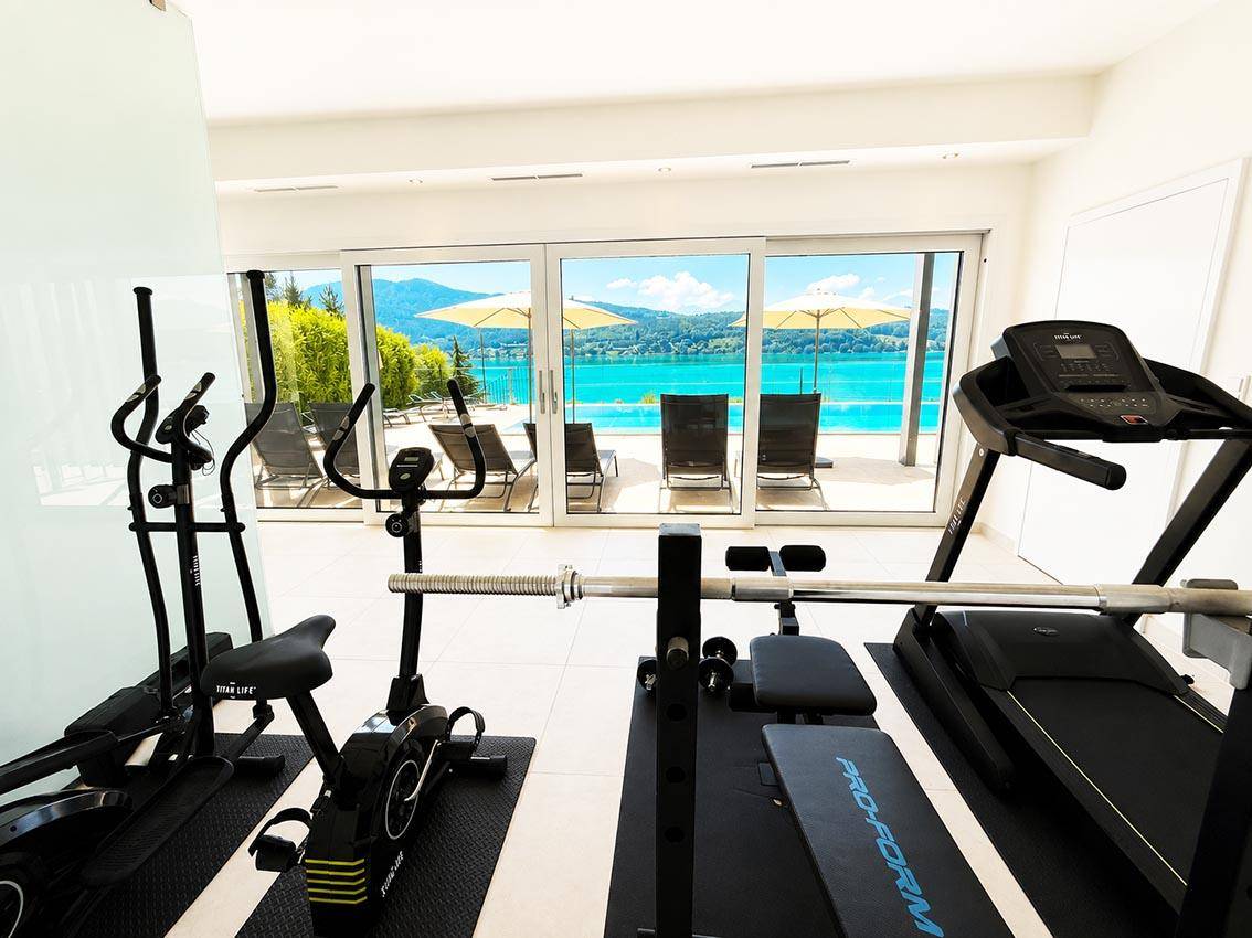 Fitnessraum mit verschiedenen Trainingsgeräten und großen Fenstern mit Blick auf den See.