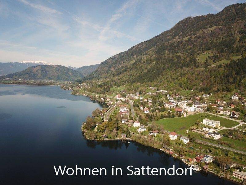 Weitläufige Luftaufnahme der Ortschaft Sattendorf am Seeufer, umgeben von bewaldeten Bergen.