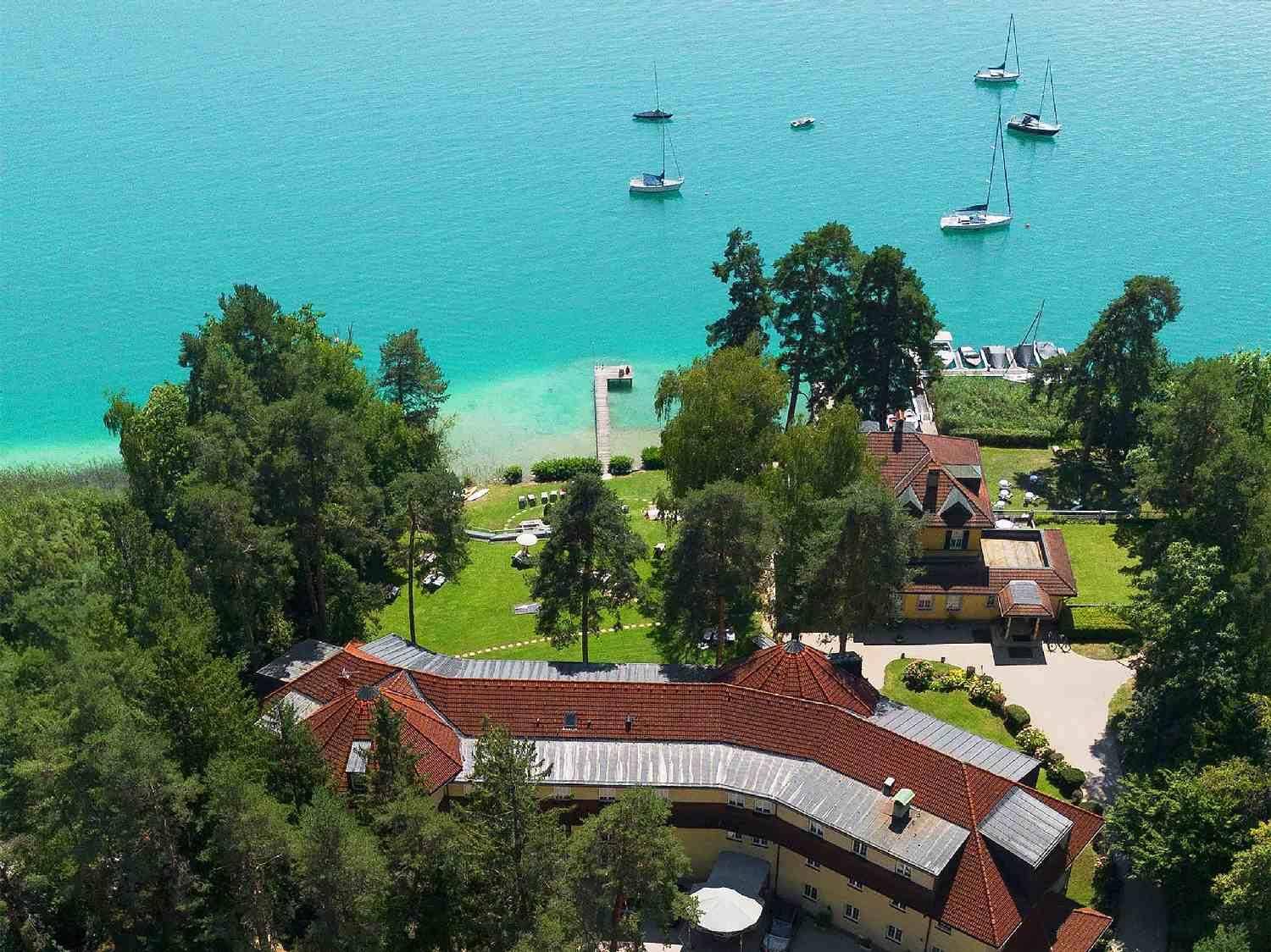 Luftaufnahme eines weitläufigen Anwesens am See mit Gebäuden, grünen Flächen und Bootsstegen.